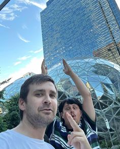 a man and woman standing in front of a glass building with their hands up to the sky