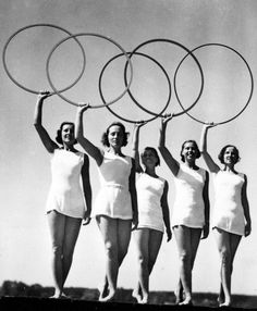 four women in white dresses hold up three large hoop rings above their heads as if they were holding tennis racquets