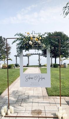 a white sign with flowers on it sitting in front of some trees and grass, surrounded by candles