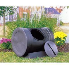 a black trash can sitting in the grass next to some flowers and plants with a house in the background