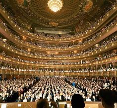 an auditorium filled with lots of people sitting on the floor and standing in front of them