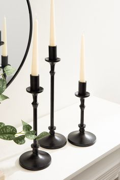 three black candles are sitting on a white table next to a mirror and plant with green leaves