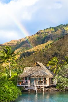 a hut on the water with a rainbow in the sky above it and palm trees