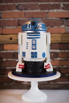 a star wars robot cake sitting on top of a white cake plate in front of a brick wall