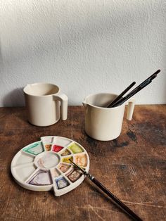 two mugs with paint and brushes sitting on a wooden table next to each other