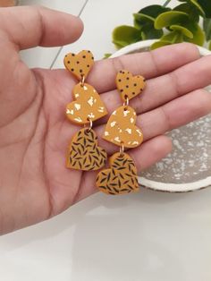 a person holding two pairs of yellow and brown earrings in their hand next to a potted plant