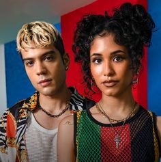 two people standing next to each other in front of a colorful wall and one person wearing a cross necklace