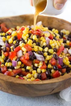 a person pouring dressing into a wooden bowl filled with corn, tomatoes and black beans