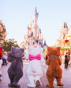 three stuffed animals are standing in front of the castle at disneyland world, one is wearing a pink bow tie