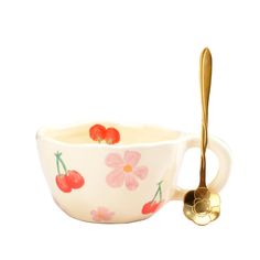 a white bowl with cherries painted on it and a spoon in the middle that is gold