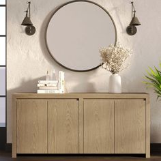 a large round mirror hanging on the wall above a wooden cabinet and vase with flowers