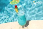 a blue drink sitting on top of a table next to a pool with an umbrella