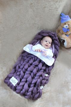 a baby wrapped in a purple blanket next to a teddy bear on a tan background