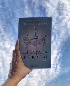 a hand holding up a book in front of a cloudy blue sky with white clouds