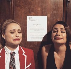 two women sitting next to each other in front of a wooden paneled wall with writing on it