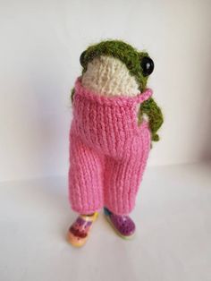 a knitted frog in a pink sweater and slippers is posed on a white surface