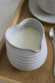 a white heart shaped bowl filled with milk next to a spoon on a wooden tray