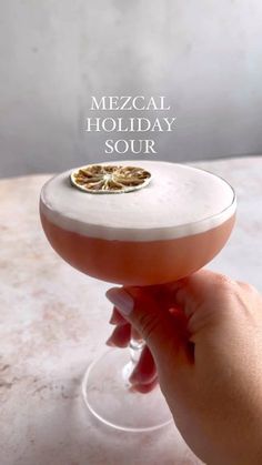 a hand holding a wine glass with a small piece of food on it that says mezcal holiday sour