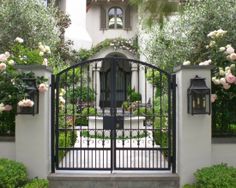 an iron gate in front of a house with roses growing on the fence and bushes around it