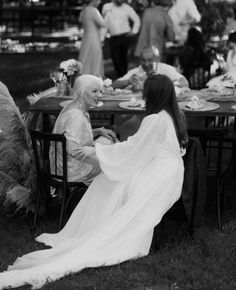 two women sitting at a table with food on it and people standing in the background