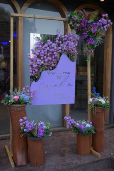 purple flowers are placed in flower pots on the sidewalk near a sign that says welcome to me