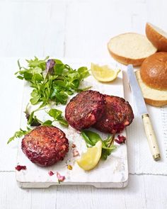 two meat patties are on a plate with bread and greens next to it, along with a knife