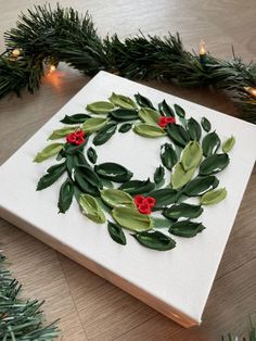 a christmas wreath made out of green leaves and red berries on a white canvas surrounded by greenery