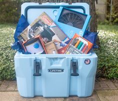 a blue cooler filled with books and magazines on top of a brick sidewalk next to a tree