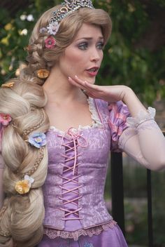 a woman with long blonde hair wearing a purple dress and tiara, holding her hand to her face