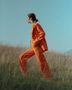 a woman in an orange outfit is walking through tall grass and looking off into the distance