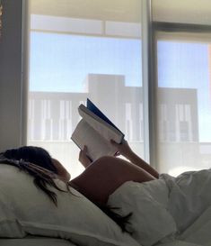 a woman laying in bed reading a book while looking out the window at the city