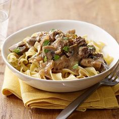 a white bowl filled with pasta and meat on top of a wooden table next to a fork