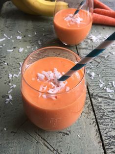 two glasses filled with orange juice on top of a wooden table next to bananas and carrots