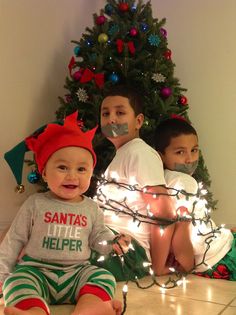 two young boys sitting on the floor in front of a christmas tree with lights around them