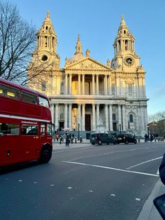 a red double decker bus driving past a tall building with two towers on it's side