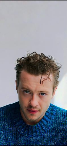 a man with curly hair wearing a blue sweater looking at the camera while holding a cell phone in his hand