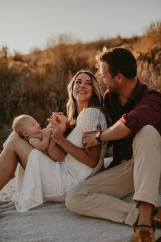 a man and woman sit on the ground with their baby in their arms as they smile at each other