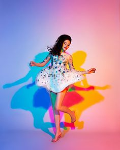 a woman in a white dress is dancing against a colorful background with her arms outstretched