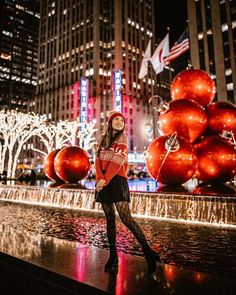 a woman standing in front of a fountain with christmas decorations on it and buildings behind her