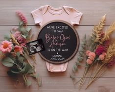 baby onesuit and flowers on a wooden surface with the words, we're excited to sasa being out there is on the way coming