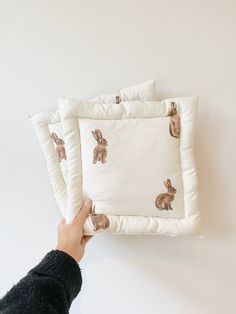 a person holding up two pillows with rabbits on them, one is white and the other is beige