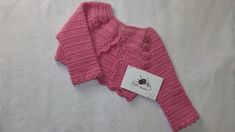 a pink knitted sweater sitting on top of a white bed next to a tag