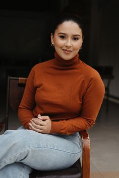 a woman sitting on top of a wooden chair wearing a brown turtle neck sweater and jeans