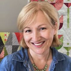 a woman with blonde hair wearing a denim shirt and smiling at the camera while standing in front of a quilted wall