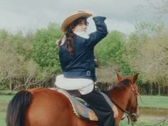 a woman riding on the back of a brown horse next to a lake and trees