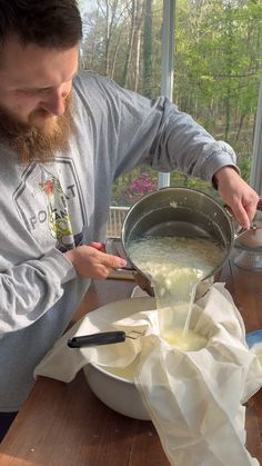 a man pouring liquid into a bowl on top of a wooden table next to a window