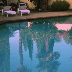 two lawn chairs sitting next to a swimming pool with palm trees reflected in the water