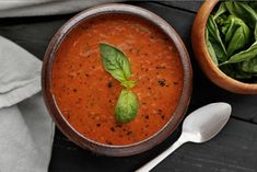 a bowl of tomato soup next to a wooden bowl filled with spinach and basil leaves