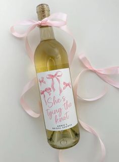 a bottle of wine with a pink ribbon around it on a white surface, and the label says she's tying the knot