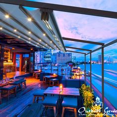 an outdoor dining area with wooden tables and chairs overlooking the water at dusk, lit by bright lights